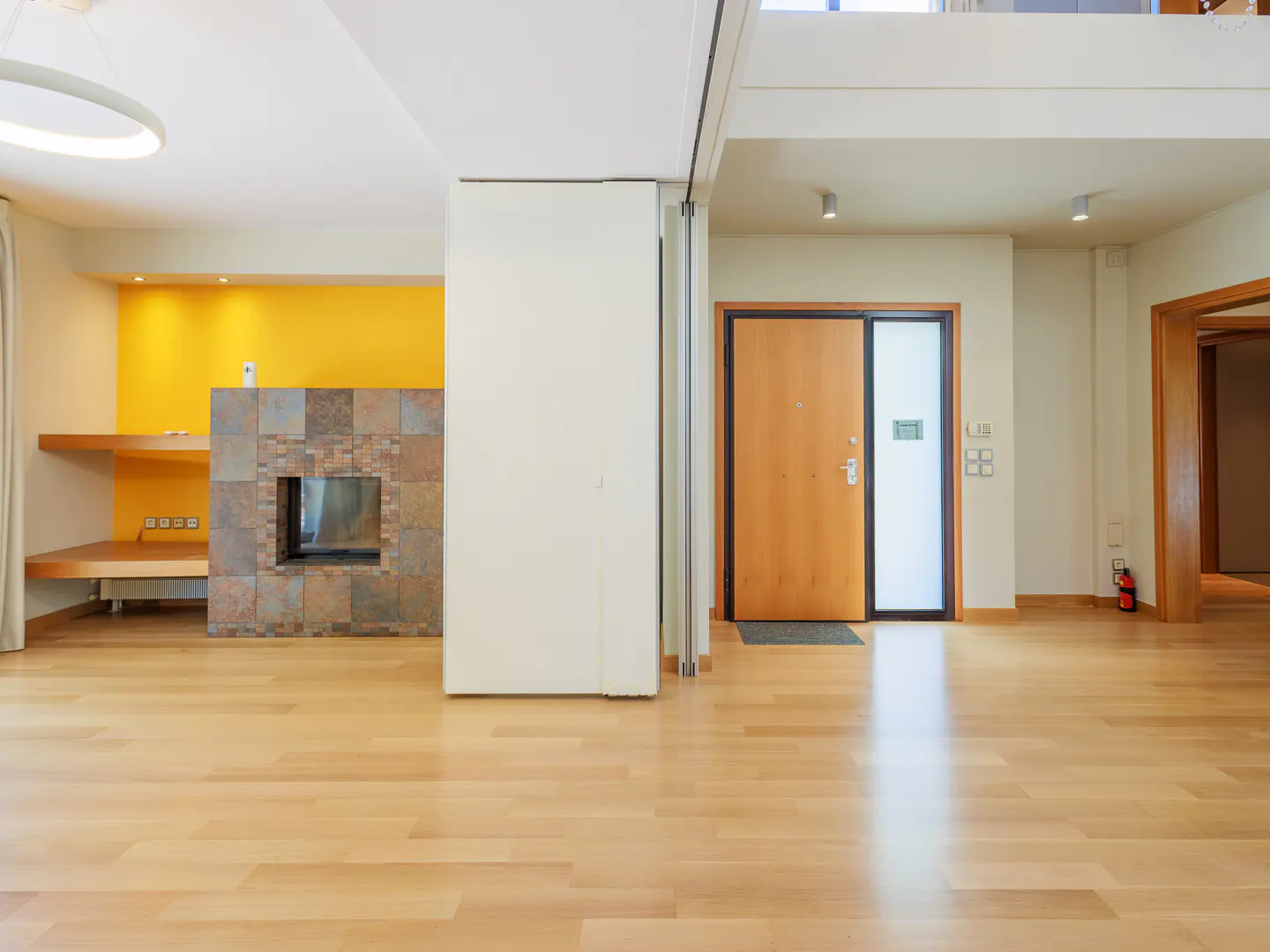 Bright, empty living room with wood floors, a stone fireplace, and yellow accent wall. A wood door is visible in the background.
