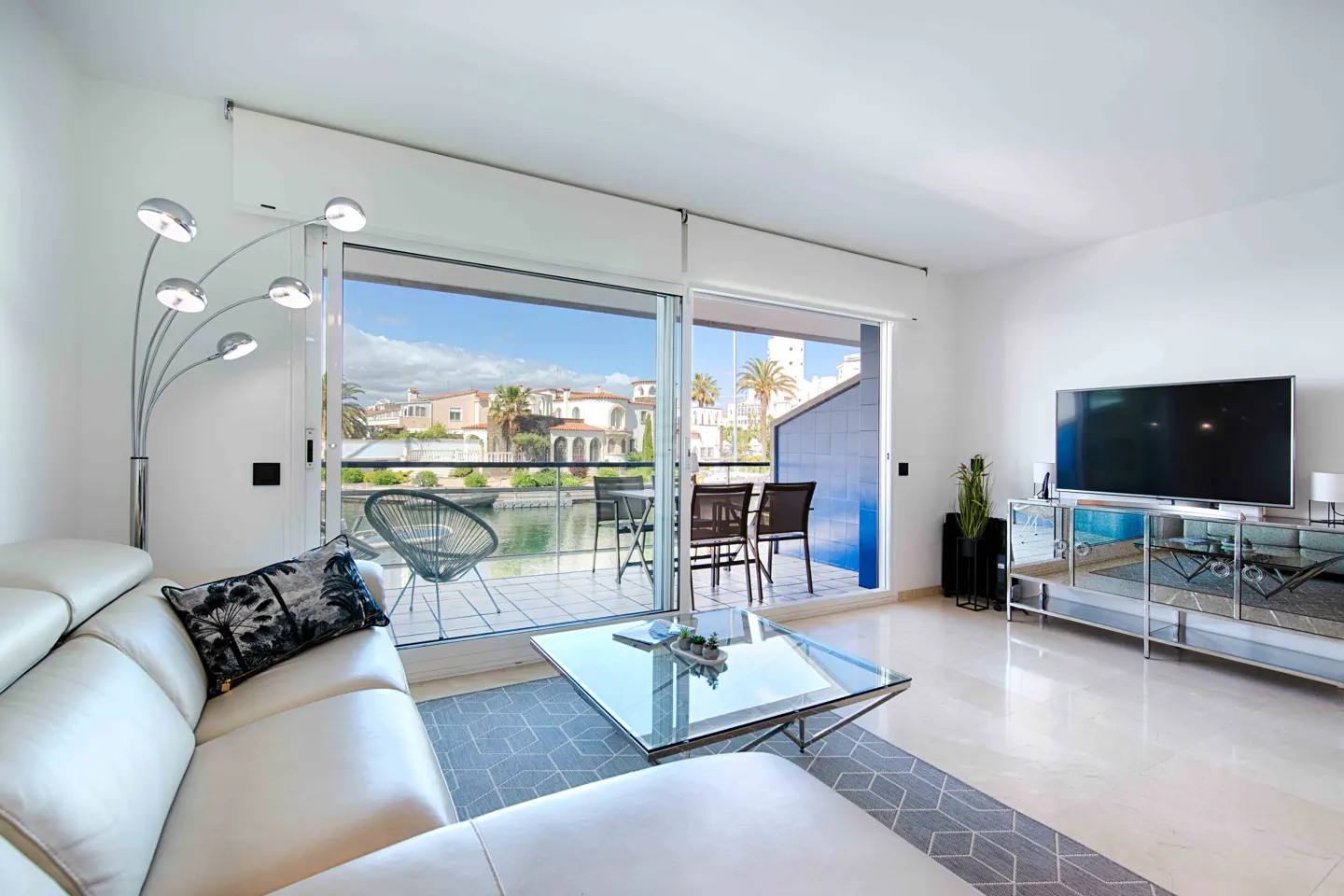 Bright living room with white sofa, glass table, and balcony overlooking a canal with buildings and palm trees. A TV sits on a mirrored console.