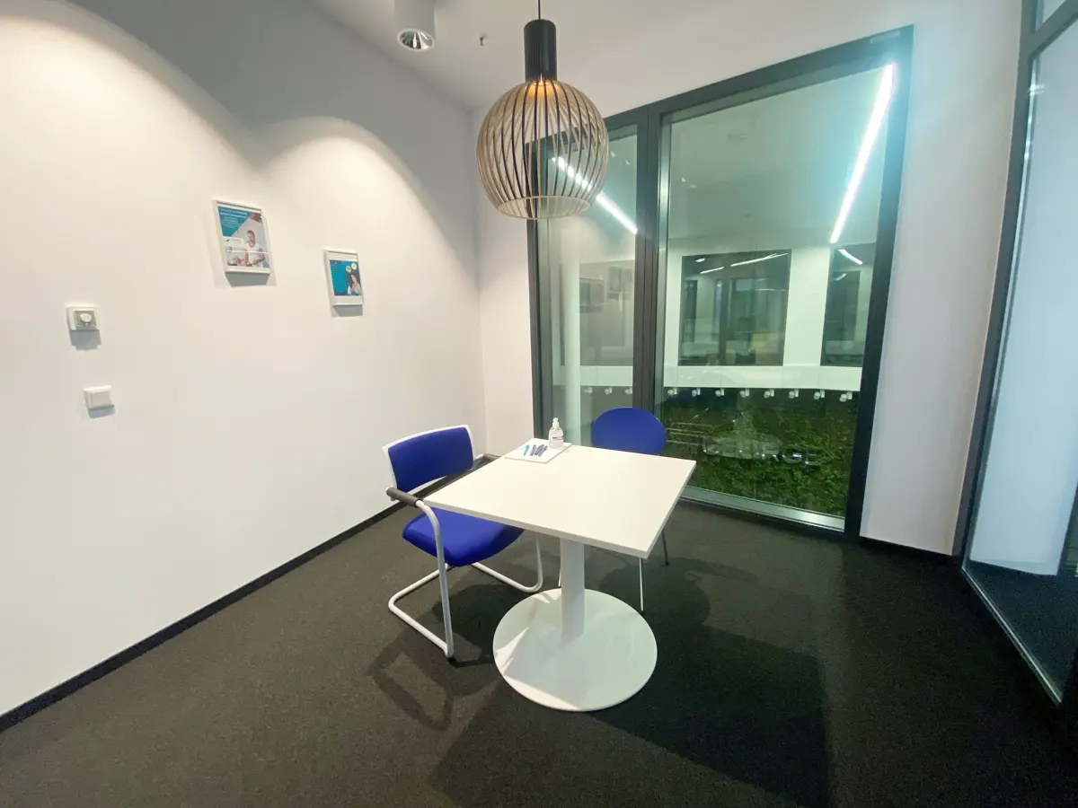 A small meeting room with a white table, two blue chairs, and a modern light fixture. A large window overlooks greenery.