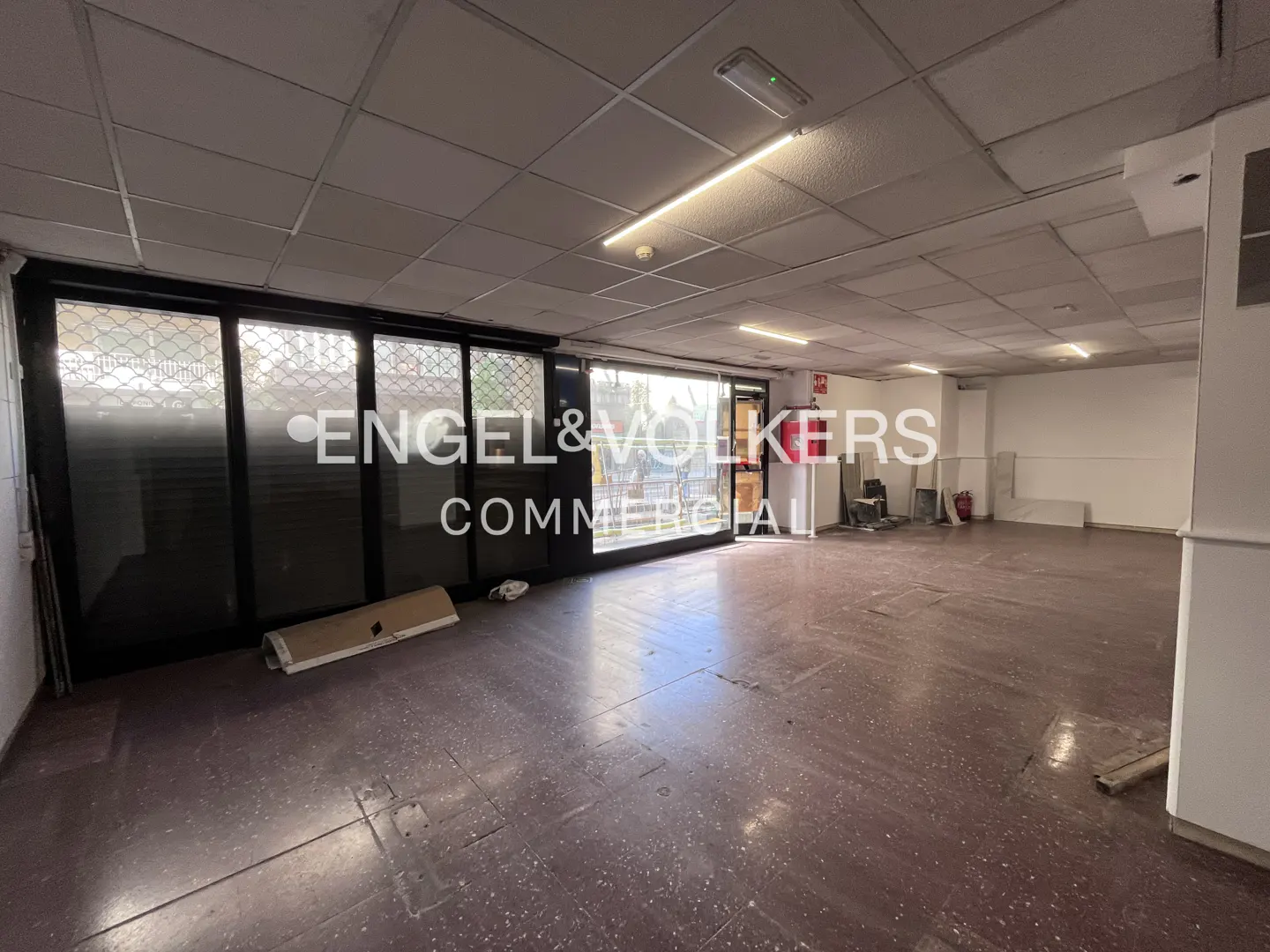 Empty commercial space with a brown floor, white walls, and a drop ceiling with fluorescent lights. A black-framed glass storefront is on the left.