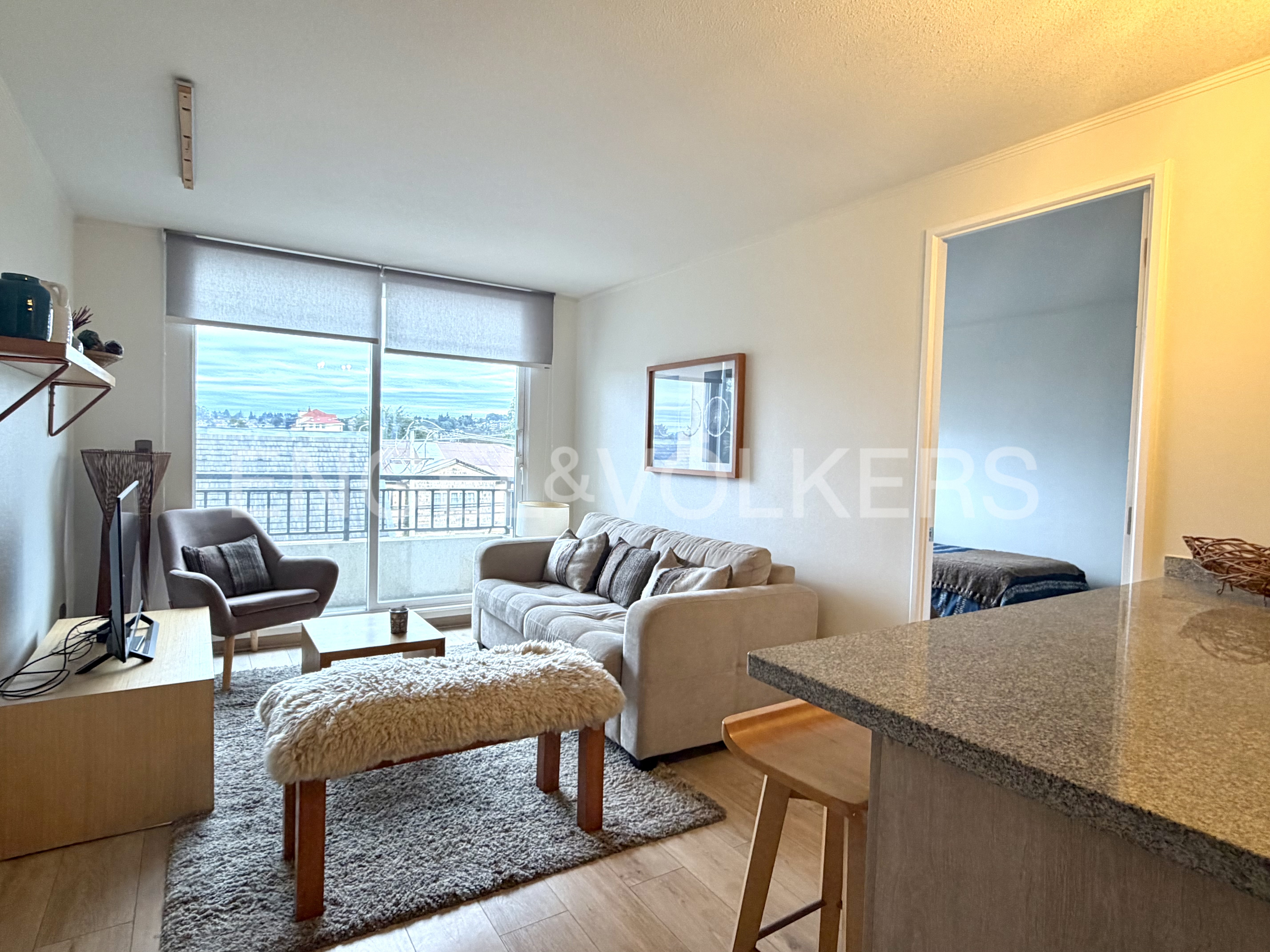 Bright living room with a beige sofa, fuzzy bench, grey armchair, and a balcony view, seen from a granite kitchen counter. [AI-generated text]
