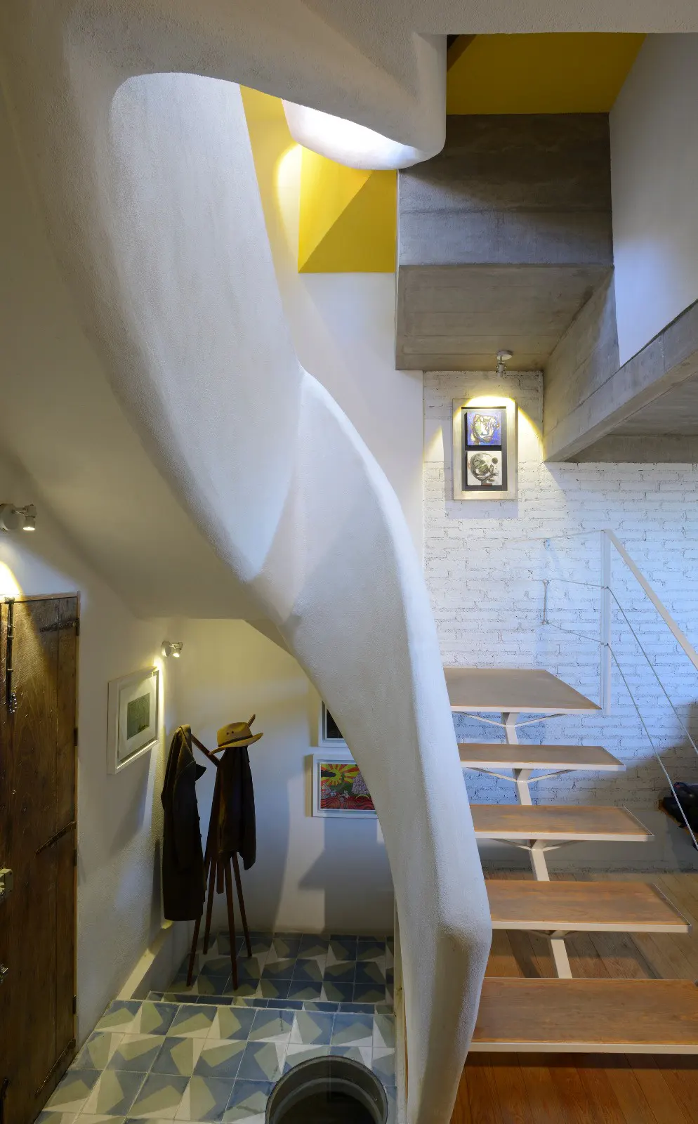 Interior view of a modern home with a white curved staircase, wooden steps, and blue patterned tile flooring. A coat rack and artwork are visible.