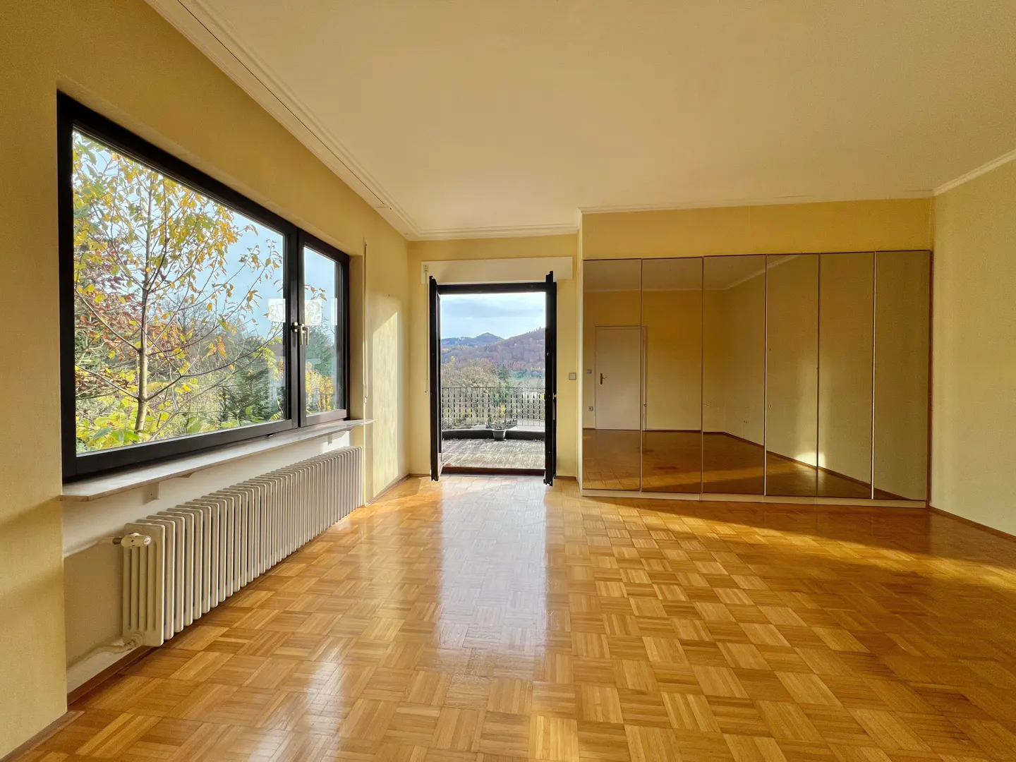 A bright, empty room with parquet floors, yellow walls, a large window with a view, and mirrored closet doors.