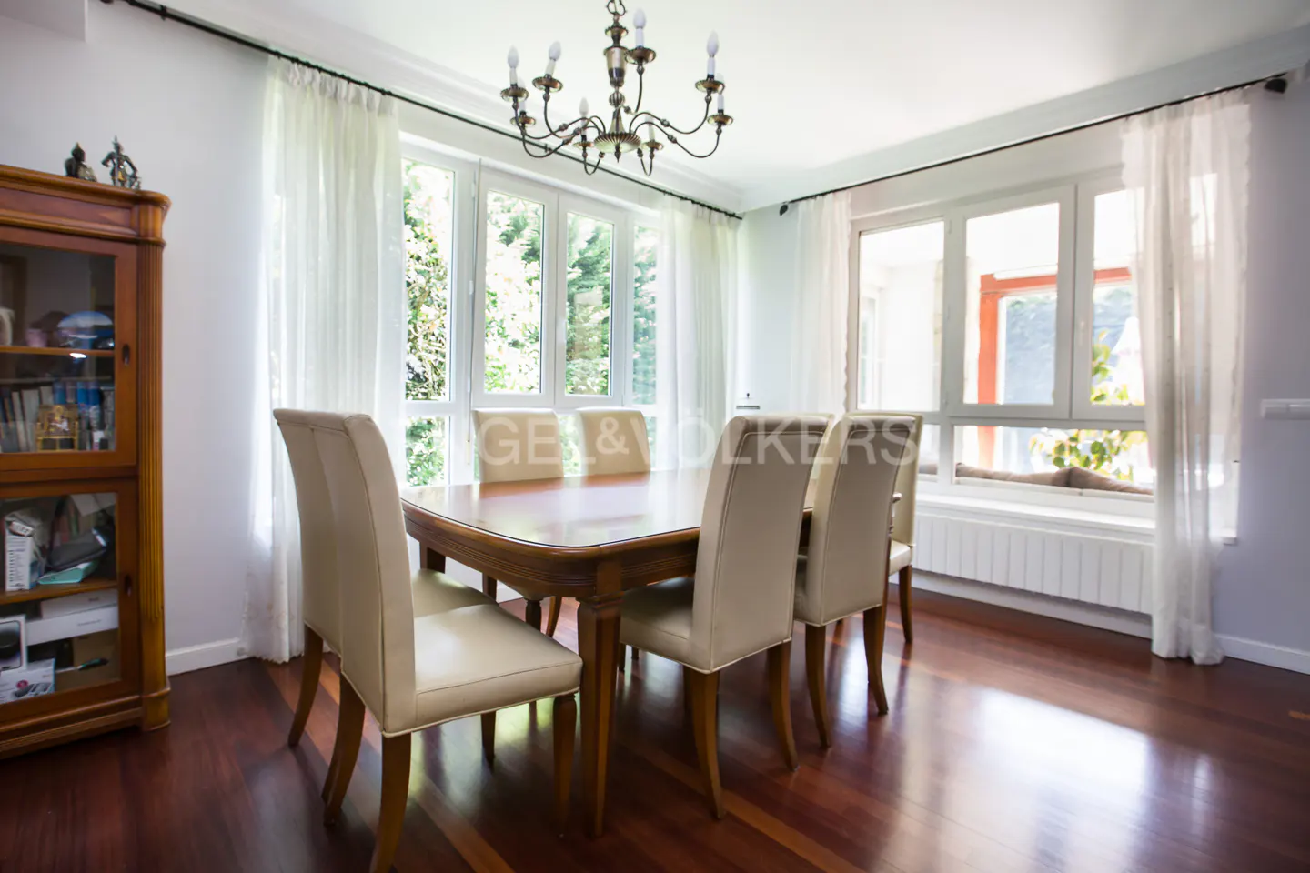 Bright dining room with a wood table, six chairs, chandelier, and large windows with sheer white curtains.