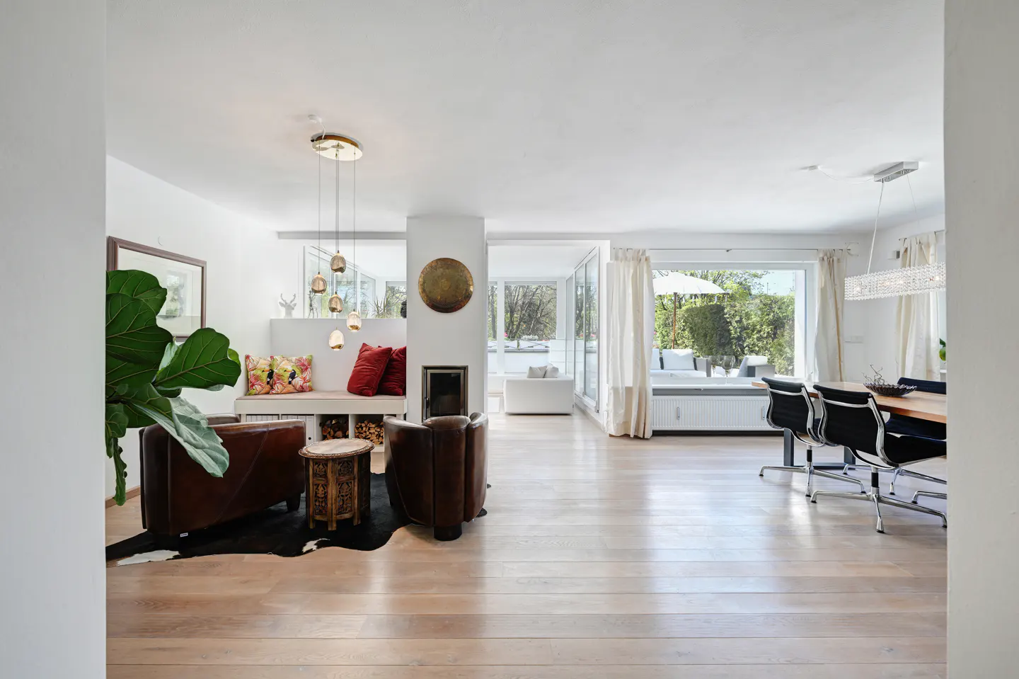 Bright open-plan living room with light wood floors, leather armchairs, a fireplace, and a dining area with garden views.