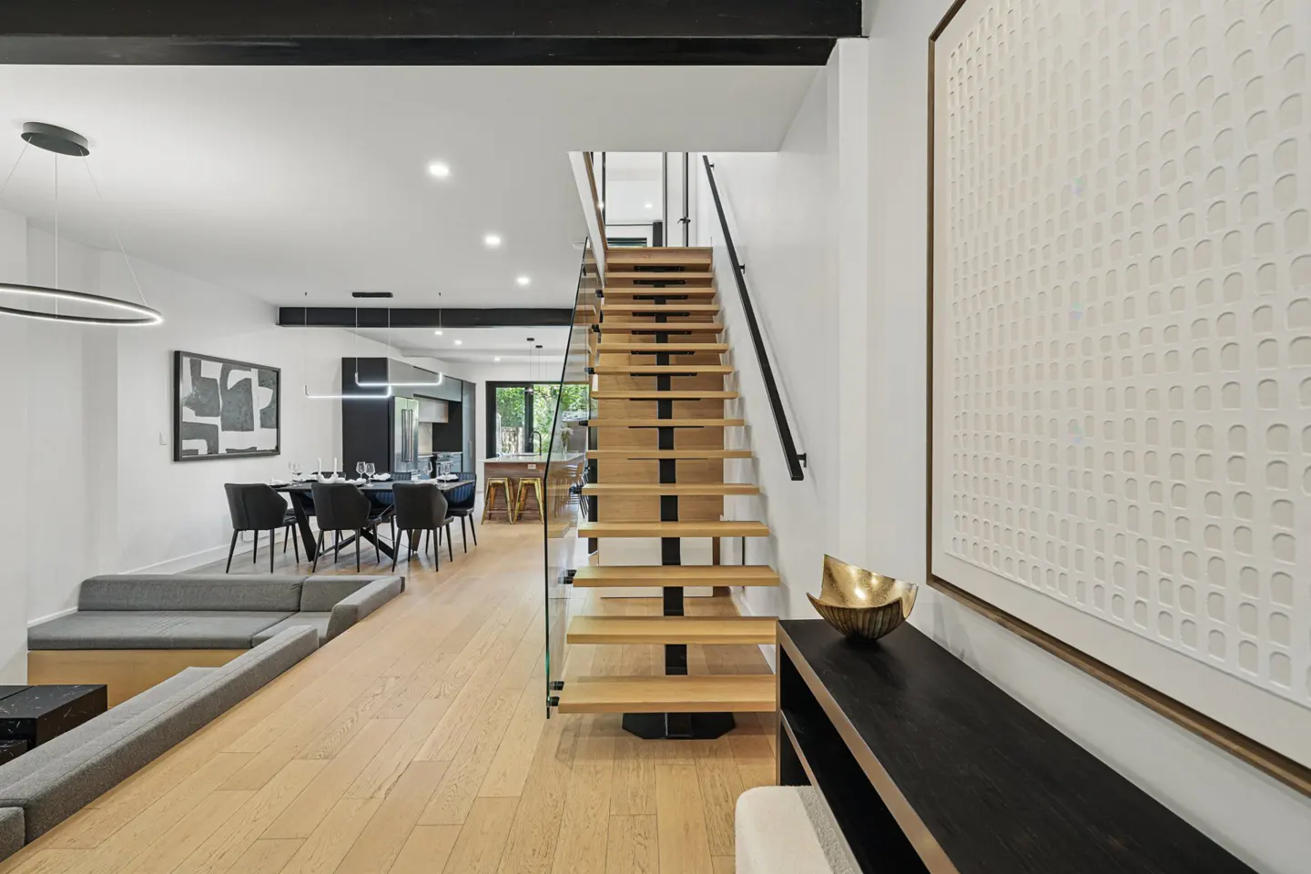 Bright, modern home interior with wood floors, open staircase, dining area with black chairs, and a gray built-in sofa. A large white textured artwork is on the wall.