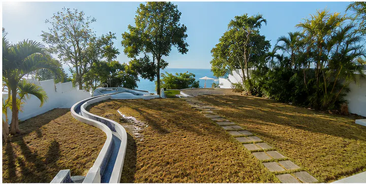 Lush green lawn with a winding water feature and stone path leading to ocean views. Trees and white walls frame the scene.