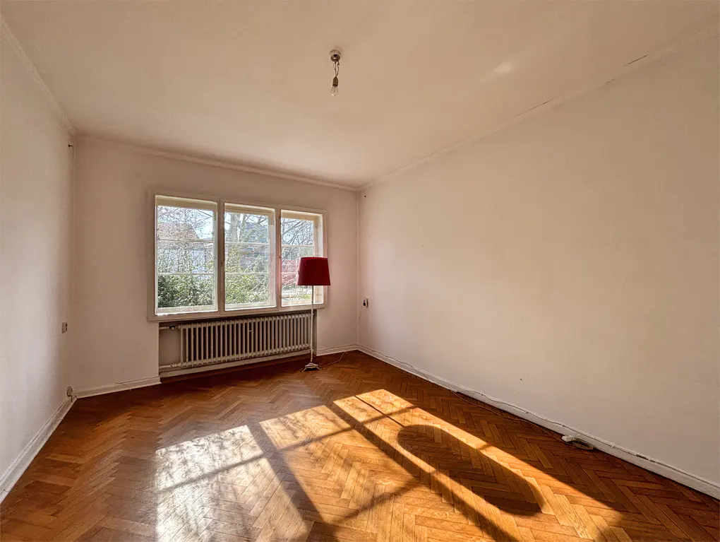 Empty room with herringbone wood floor, white walls, radiator under window, and red floor lamp. Sunlight streams in.