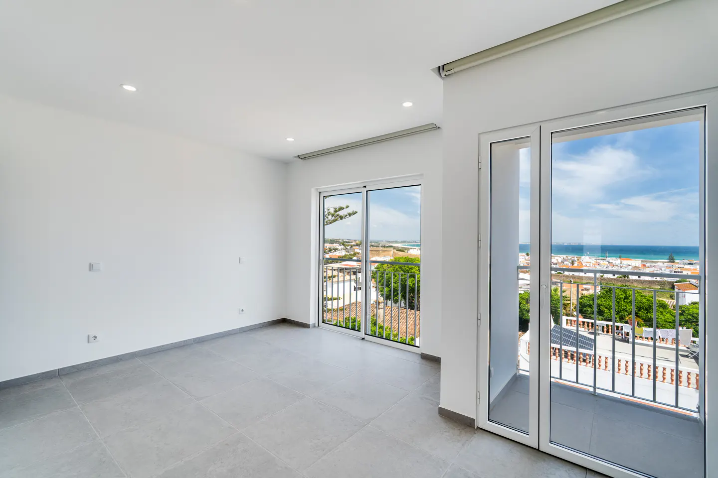 Bright, empty room with gray tile floor, white walls, and two sliding glass doors leading to balconies with ocean views.