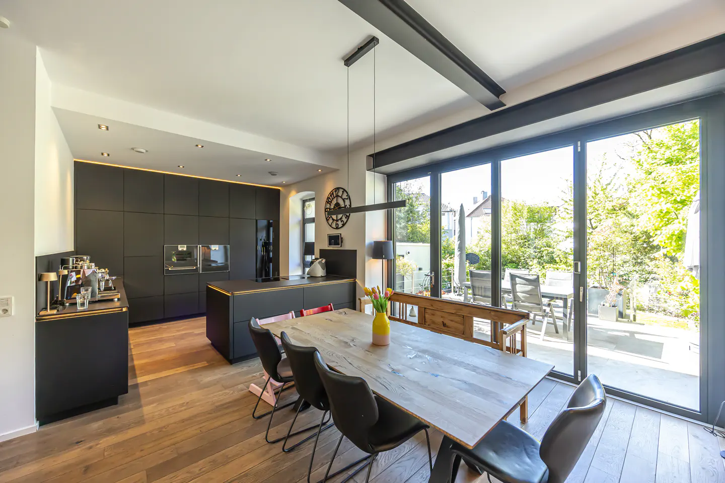 A modern kitchen with black cabinets, a wood table with black chairs, and sliding glass doors to a green backyard.