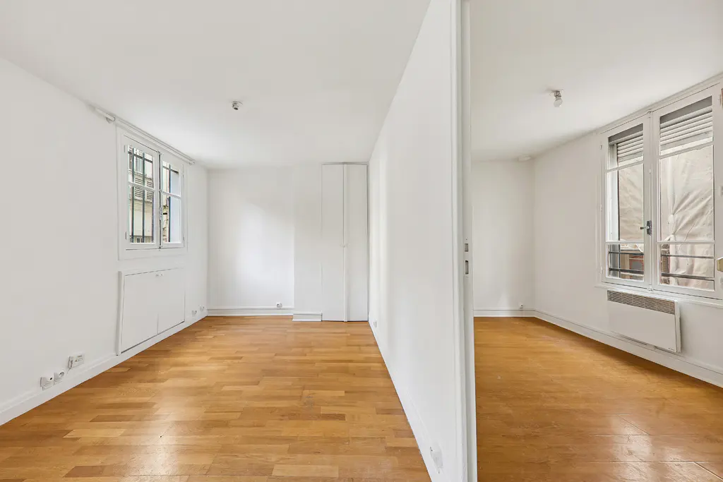 Bright, empty apartment with hardwood floors and white walls. A partial wall divides the space, with windows visible in both sections.