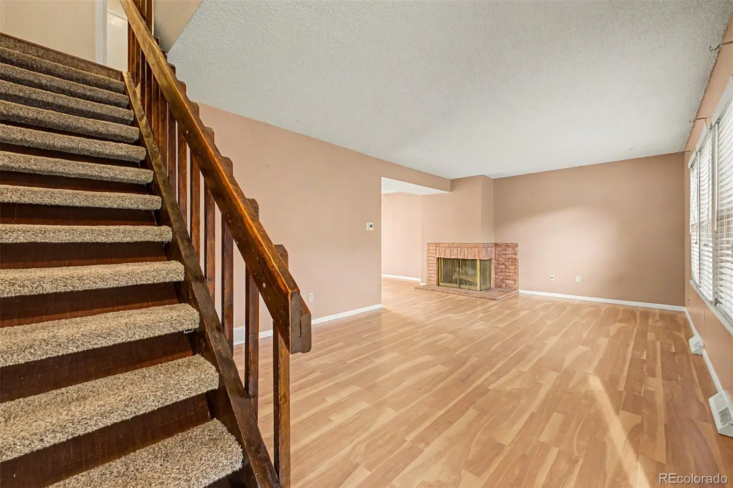 Interior view of a living room with a staircase, wood floors, a brick fireplace, and tan walls.