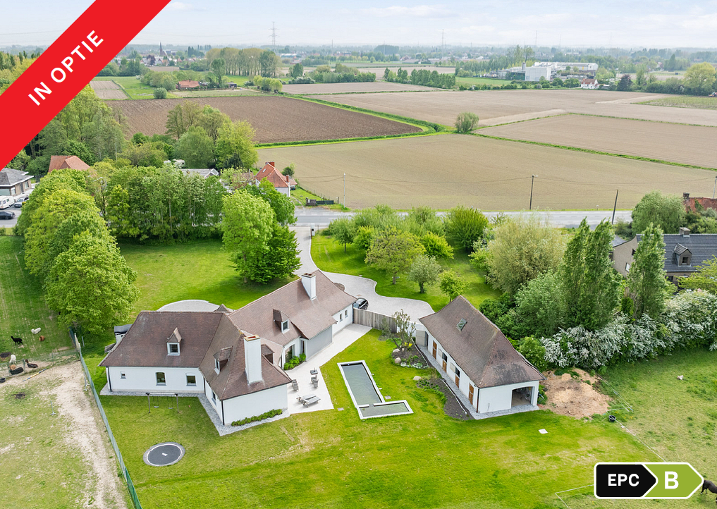 Aerial view of a white house with a brown roof, pool, and outbuilding, surrounded by green lawns and rural farmland.