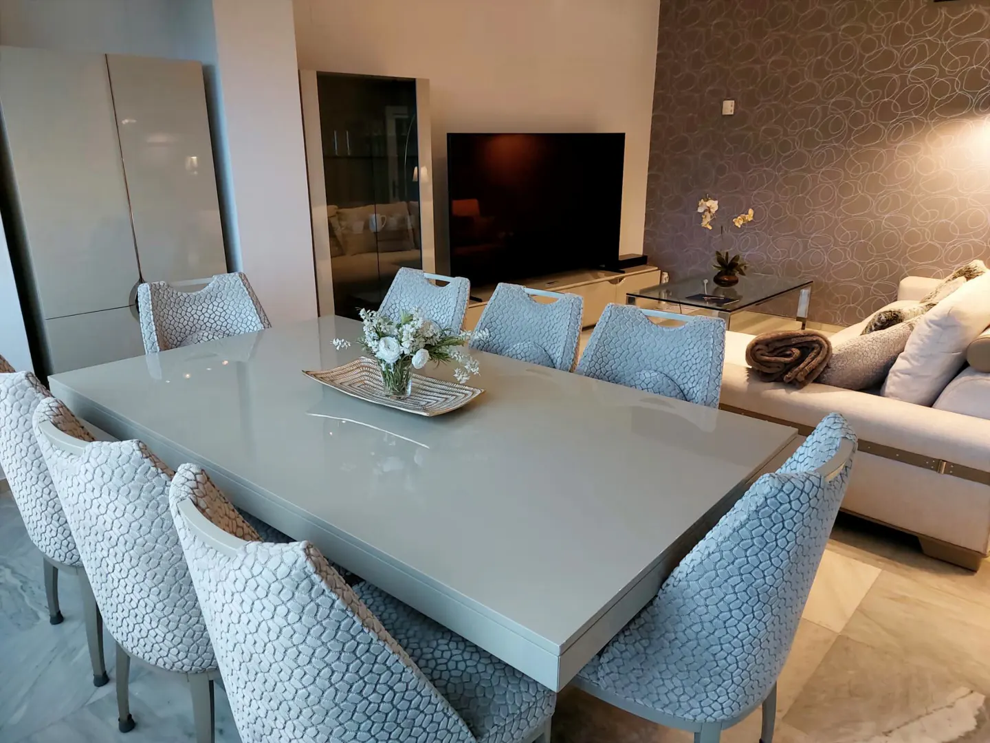 A modern dining room features a long, light gray table with textured gray chairs, a floral centerpiece, and a living area in the background.