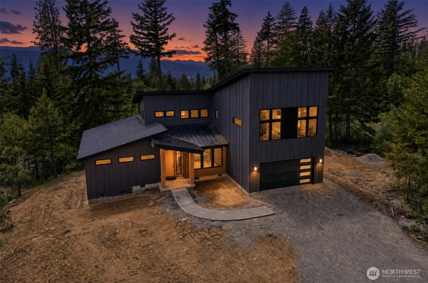 Modern gray house with black roof and garage, surrounded by trees at dusk. A concrete path leads to the front door.