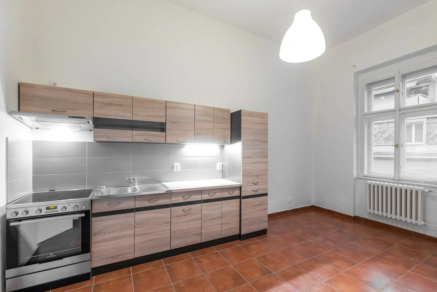 Bright kitchen with wood cabinets, stainless steel appliances, and terracotta tile floor. A window with a radiator is on the right.