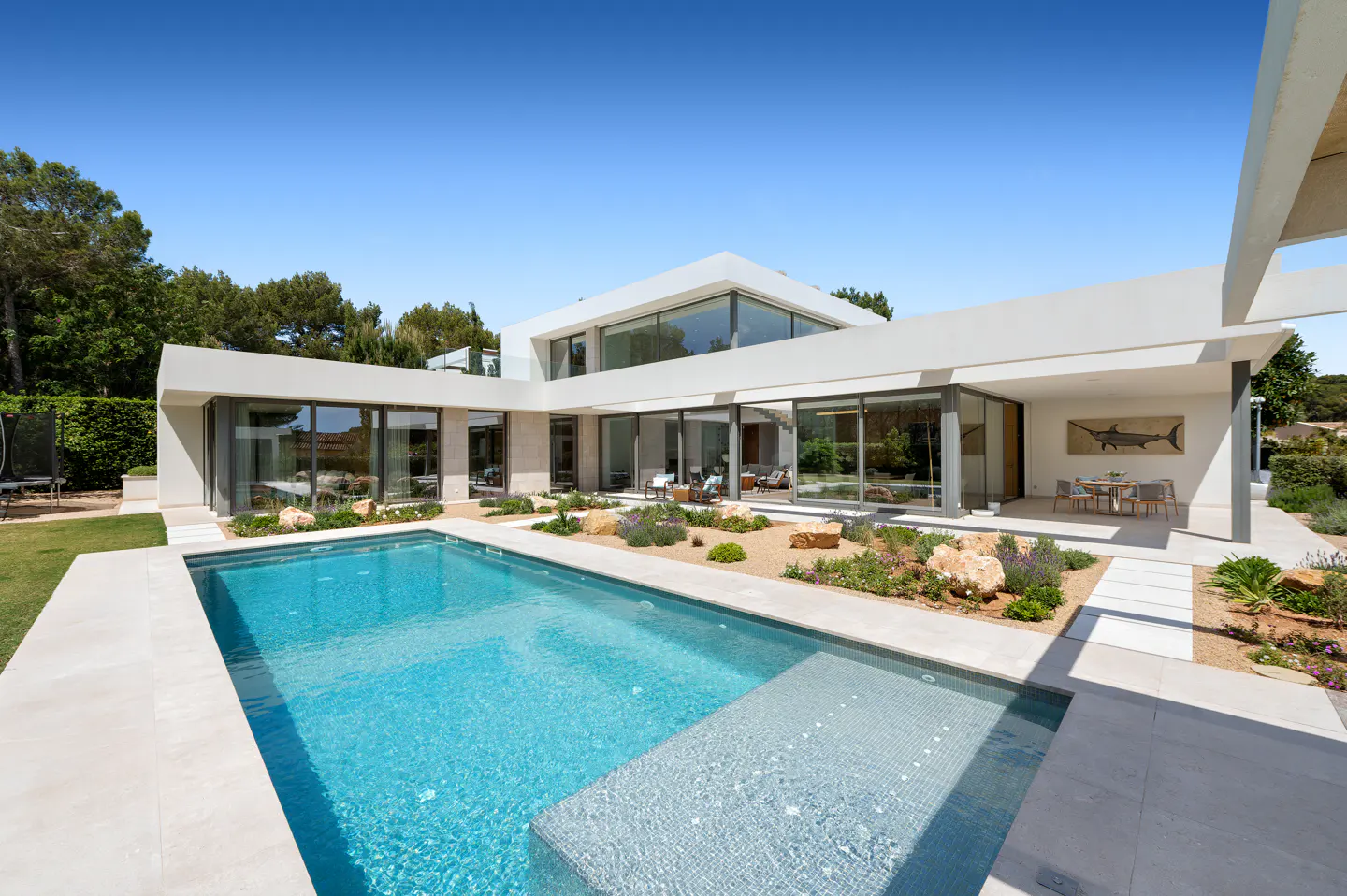 Modern white house with a blue pool in the foreground. Large glass windows reflect the interior. A patio with a table and chairs is visible.