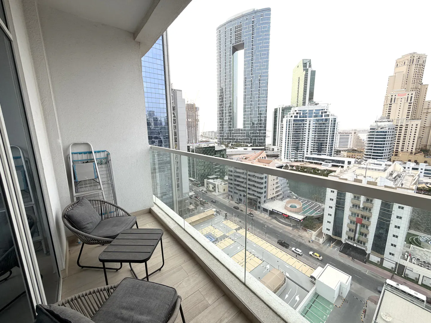 Balcony view with gray chairs, table, and clothes drying rack. Cityscape with tall buildings visible through glass railing.