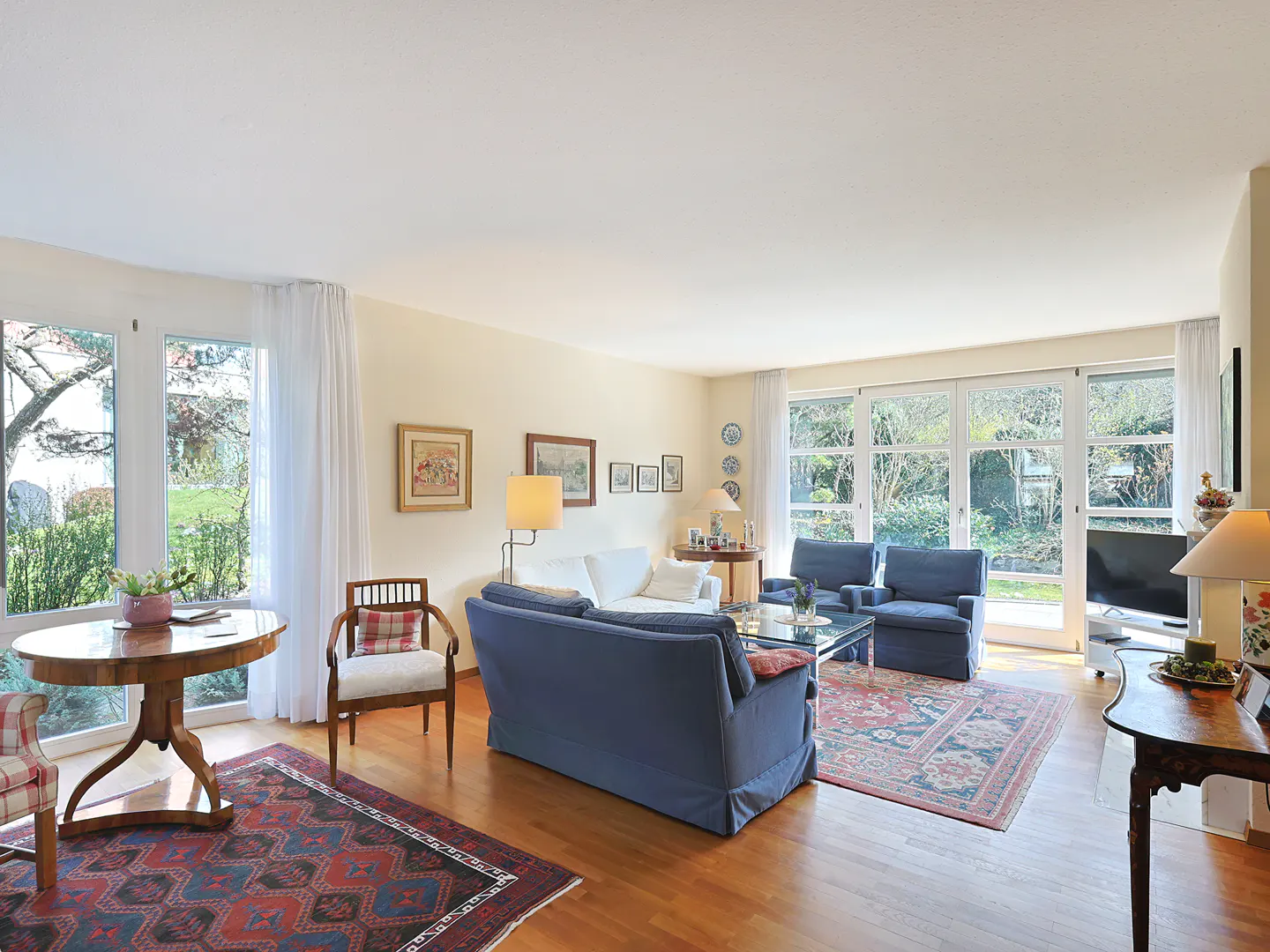 Bright living room with hardwood floors, blue sofas, and large windows overlooking a green yard. A patterned rug adds warmth.