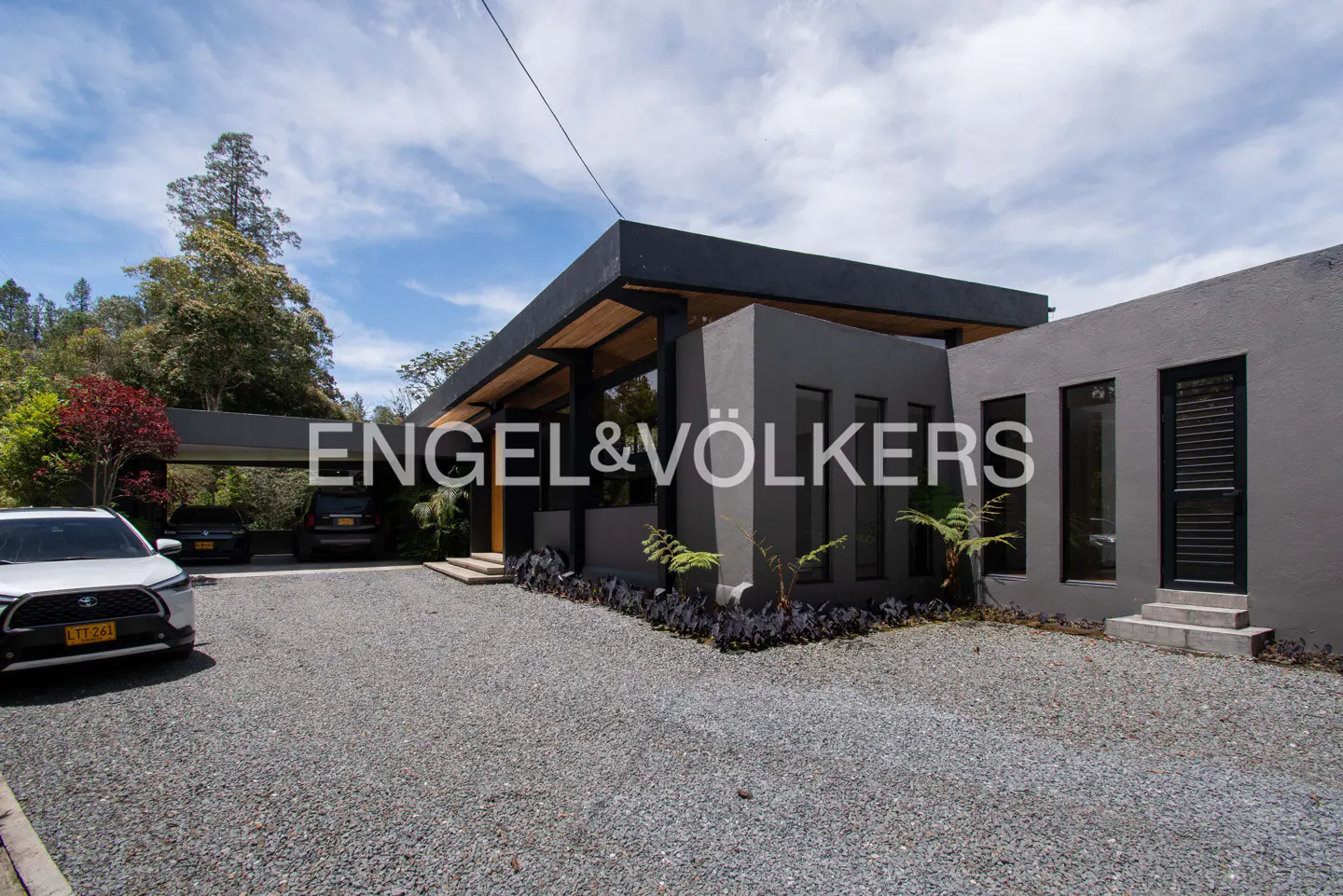 Exterior view of a modern gray house with a black roof and a gravel driveway with cars parked under a carport.