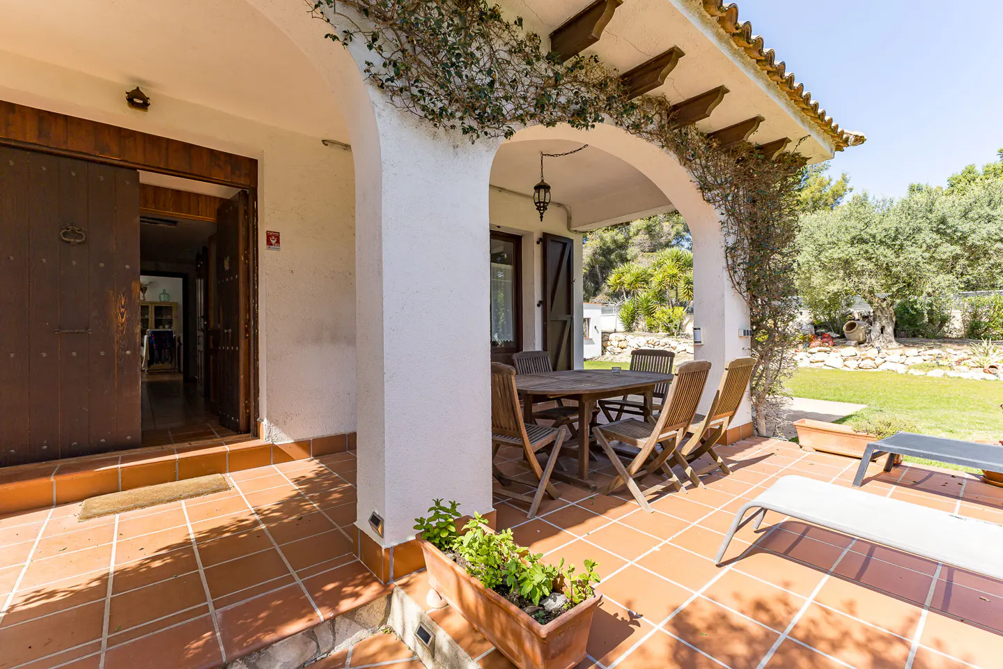 Exterior view of a Spanish-style home with a terracotta-tiled patio, wooden table and chairs, and a garden with trees.