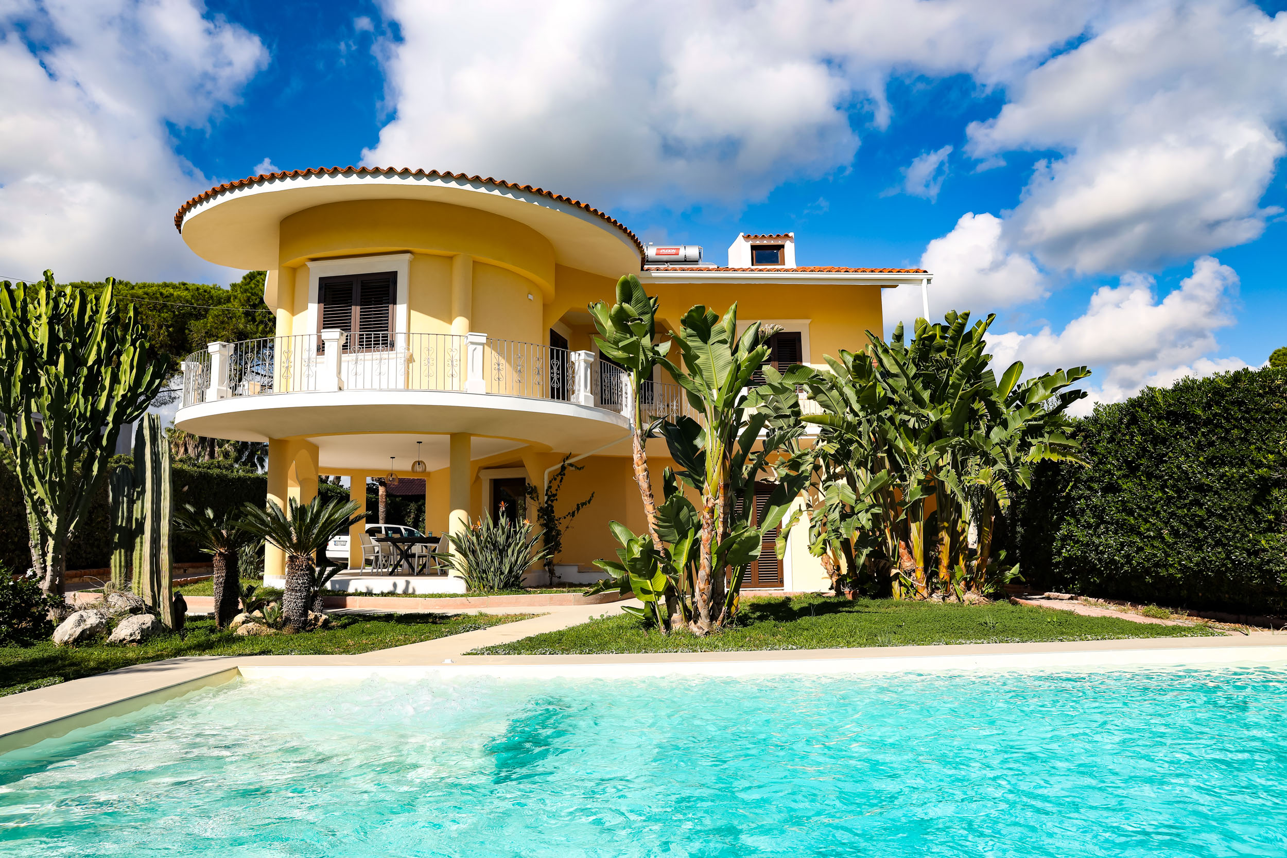 A yellow two-story house with a balcony, surrounded by lush greenery and a turquoise swimming pool.