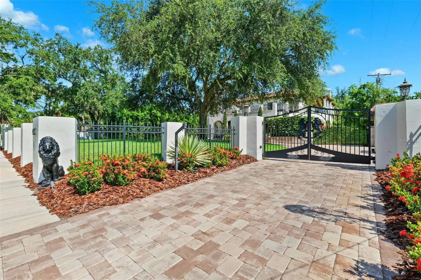 Gated driveway leading to a white house. Black iron fence and gate with lion statues. Brick pavers and green lawn.