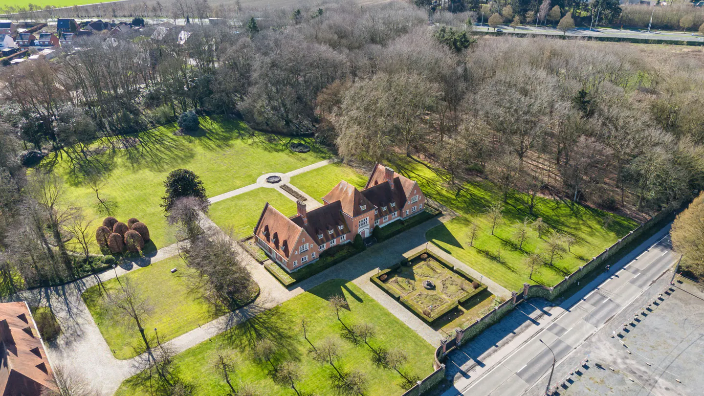 Aerial view of a large, red-roofed house surrounded by green lawns, trees, and gardens.