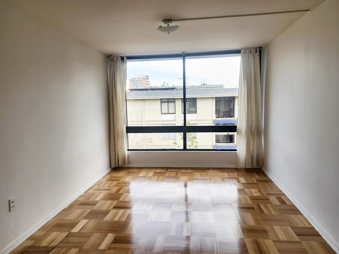 Empty room with parquet floor, white walls, and large window with white curtains overlooking a building.