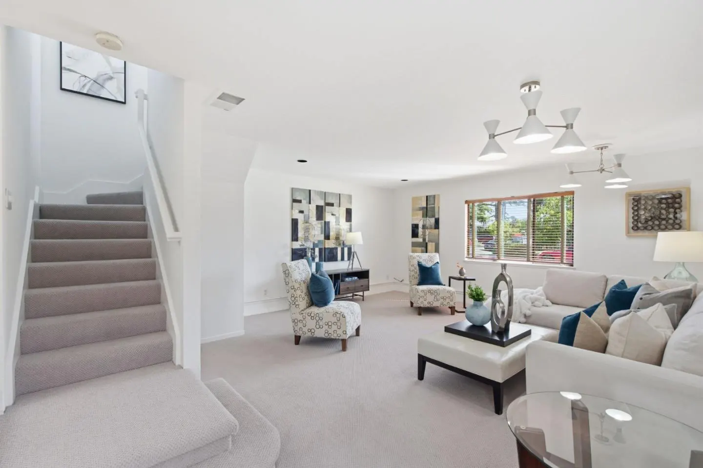 Bright living room with white walls, gray carpet, and stairs. Furniture includes a white sofa, patterned chairs, and modern lighting.