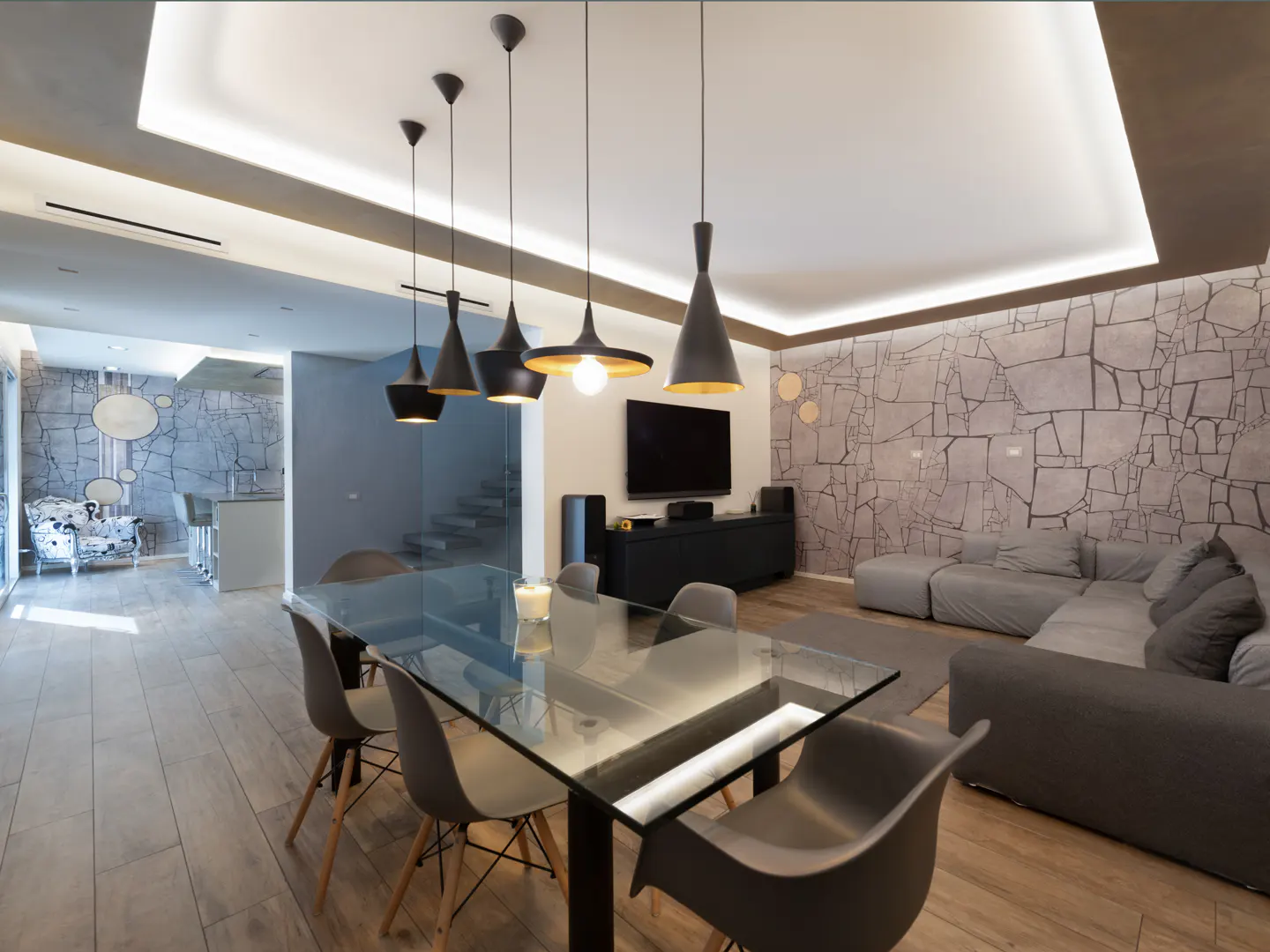 A modern living and dining area with a glass table, gray chairs, and pendant lights. A gray sofa and TV are in the background.