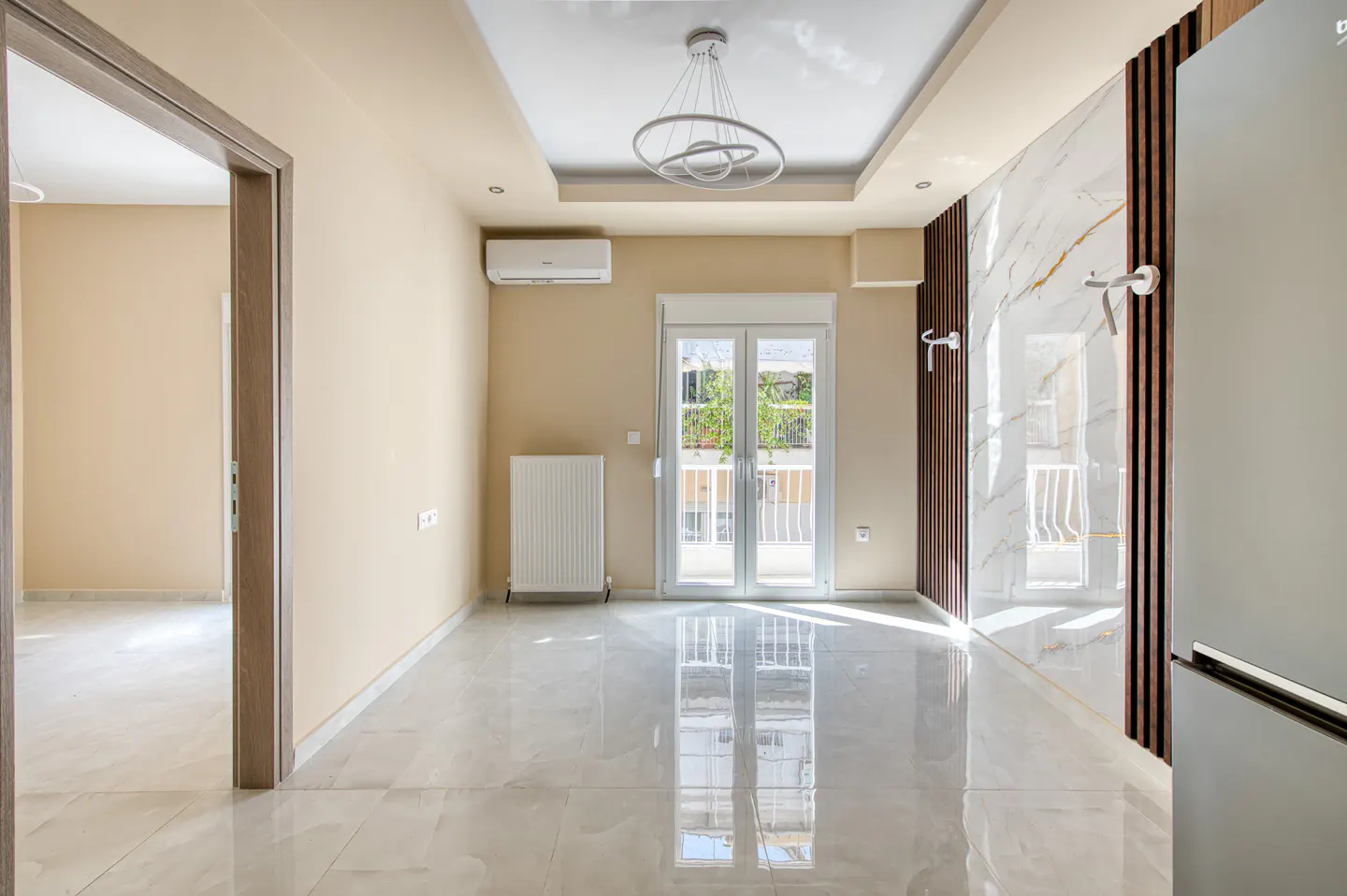 Bright, empty room with marble floors, beige walls, and a modern light fixture. French doors lead to a balcony. A refrigerator is on the right.