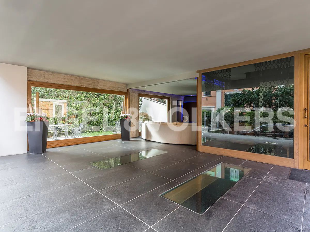 Modern foyer with dark tile floors, glass floor panels, and large windows overlooking a garden. A curved white sofa sits in the background.