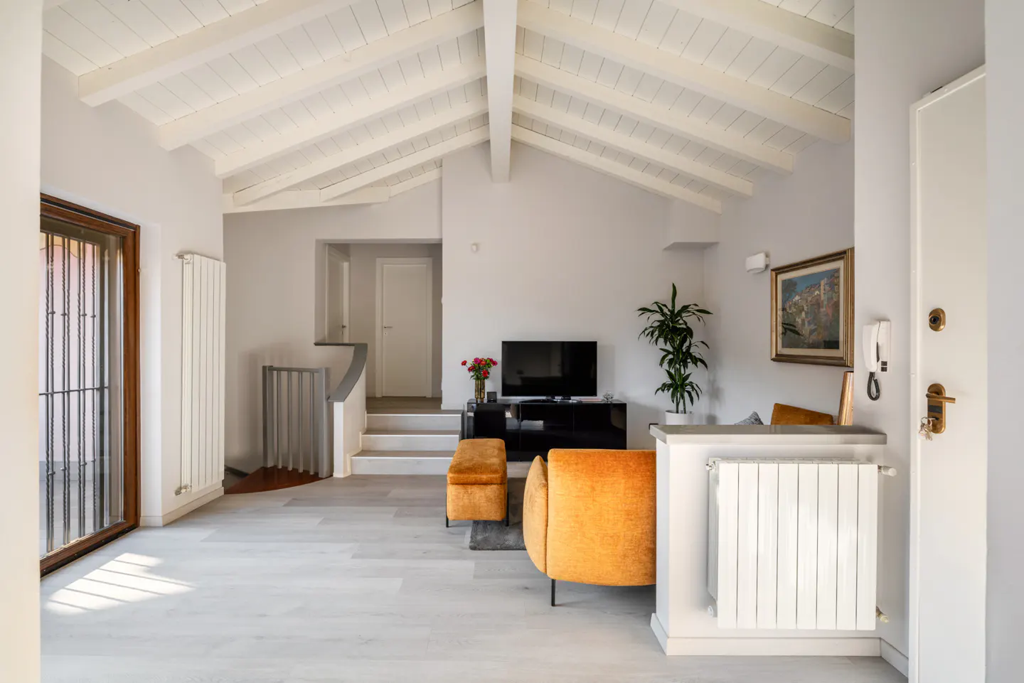 Bright living room with white vaulted ceiling, gray walls, and light wood floors. An orange sofa faces a black TV stand. Stairs lead to an upper level.