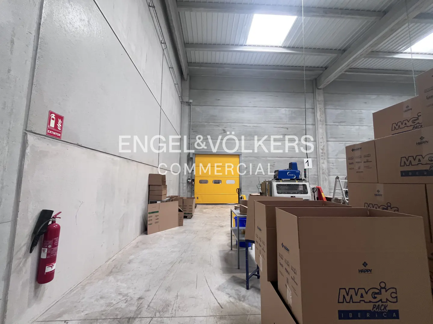 Warehouse interior with concrete walls, a yellow loading door, and stacks of cardboard boxes. A red fire extinguisher is mounted on the wall.