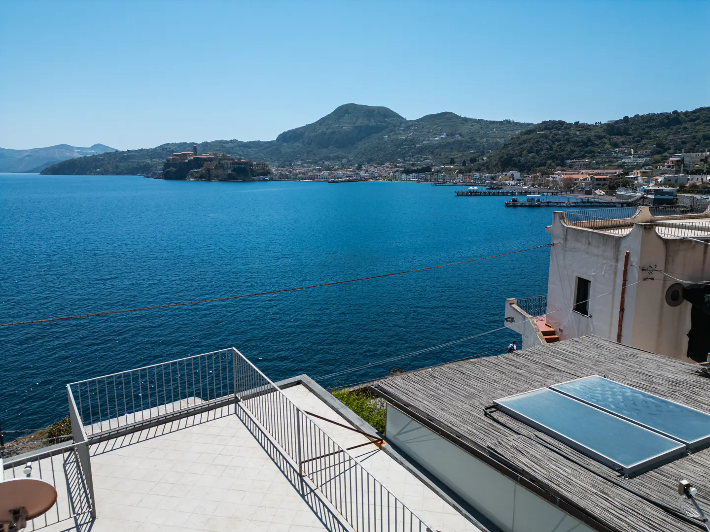 Ocean view from a rooftop terrace with white railings and solar panels. Blue water and green hills in the background.