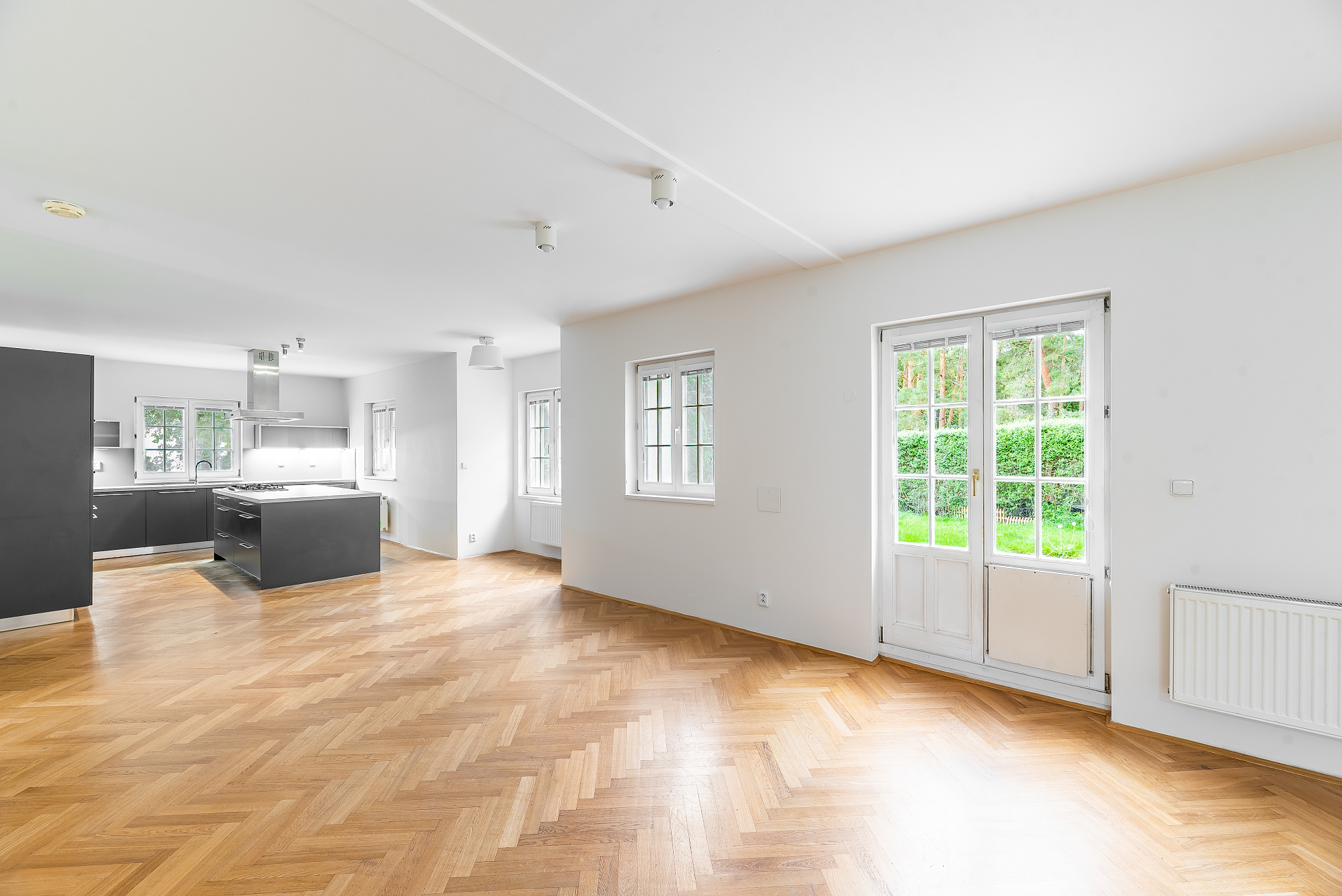 Bright open-plan living area with herringbone wood floors, white walls, and a modern kitchen with dark grey cabinets.