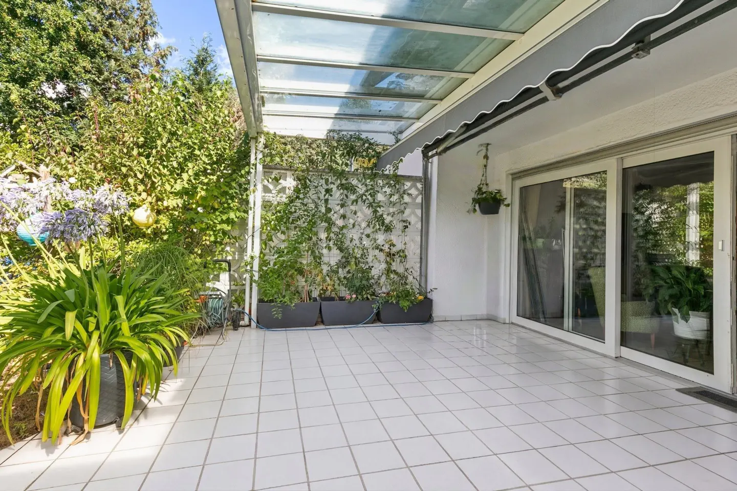 Covered patio with white tile floor, sliding glass doors, and lush greenery. A gray awning provides shade.