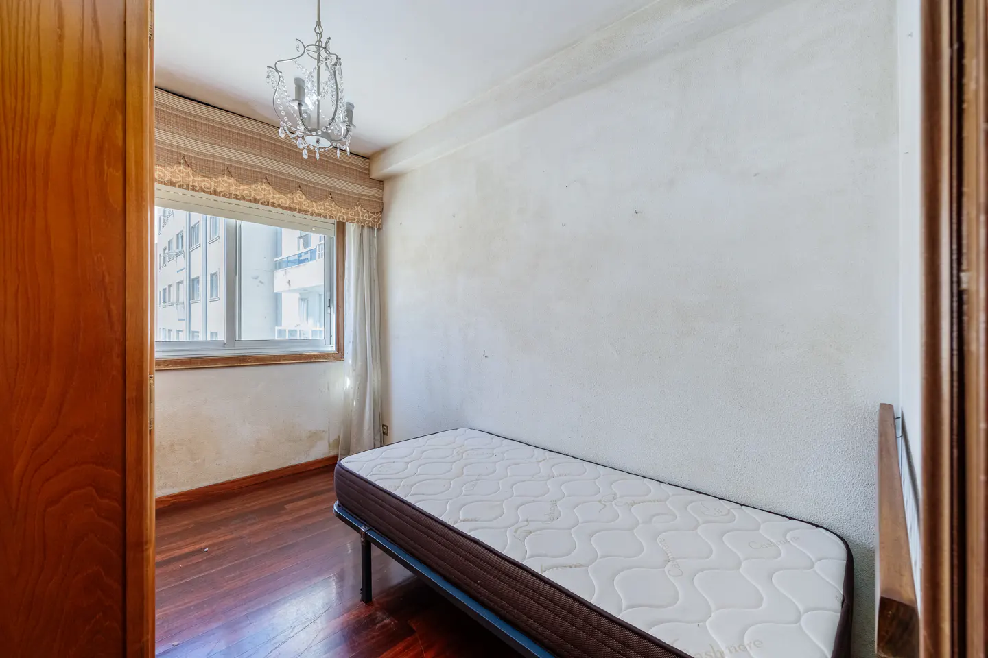 Small bedroom with dark wood floors, a single bed, a crystal chandelier, and a window with patterned valance curtains.