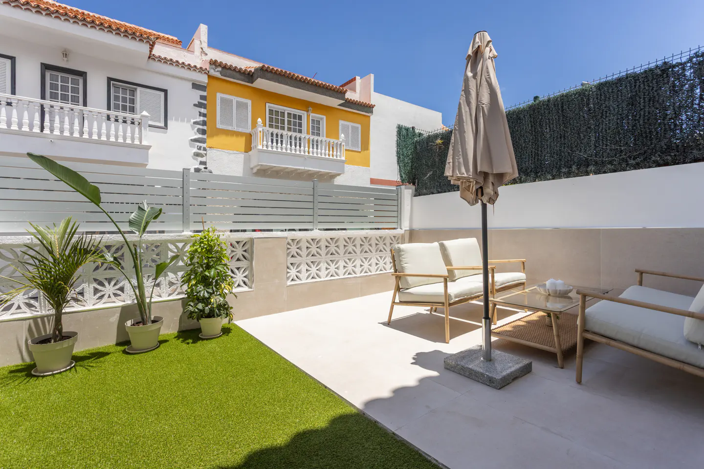 Outdoor patio with white furniture, a closed beige umbrella, and a small glass table. There is a small patch of artificial grass.