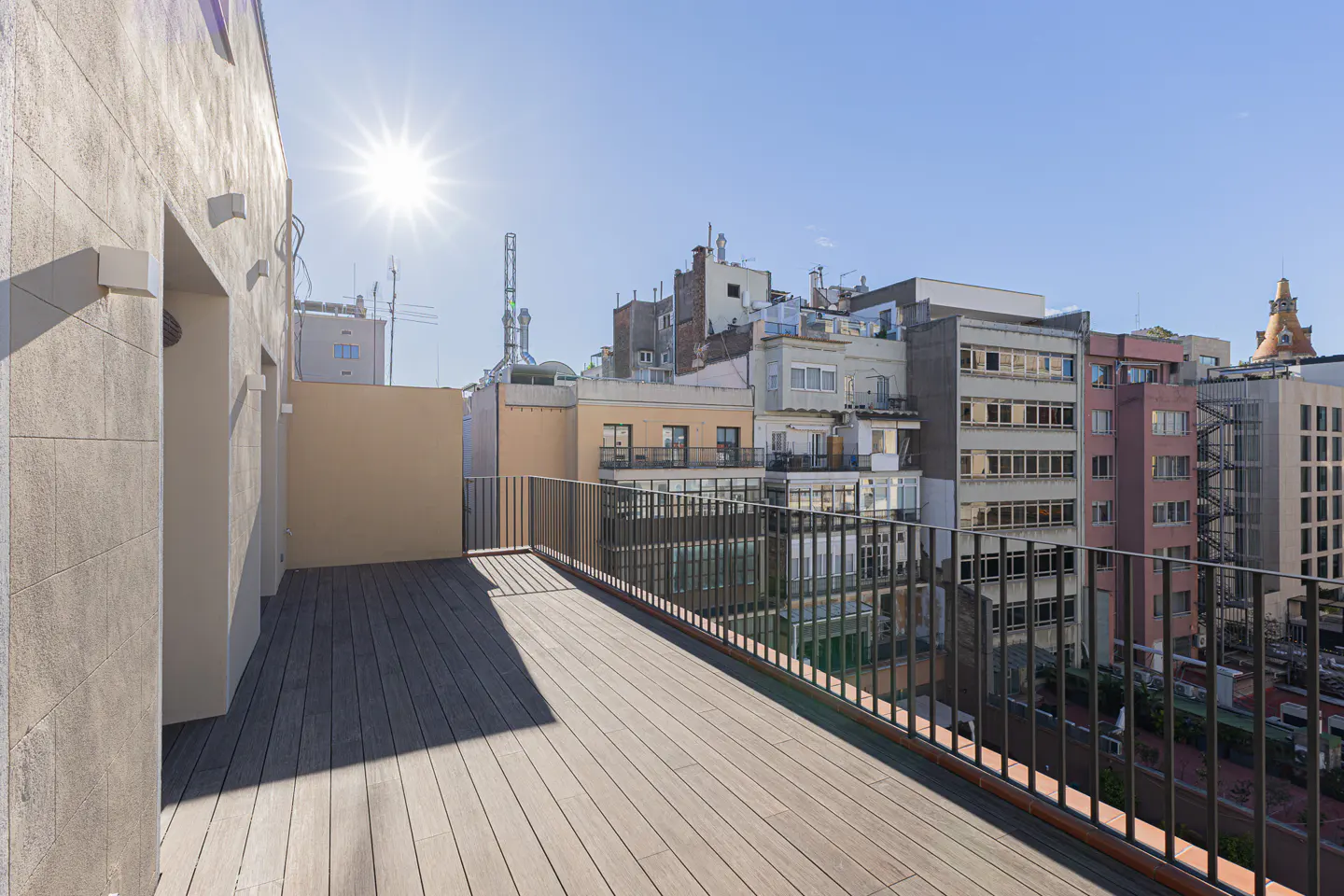A rooftop terrace with wood flooring and a metal railing overlooks a cityscape under a bright, sunny sky.