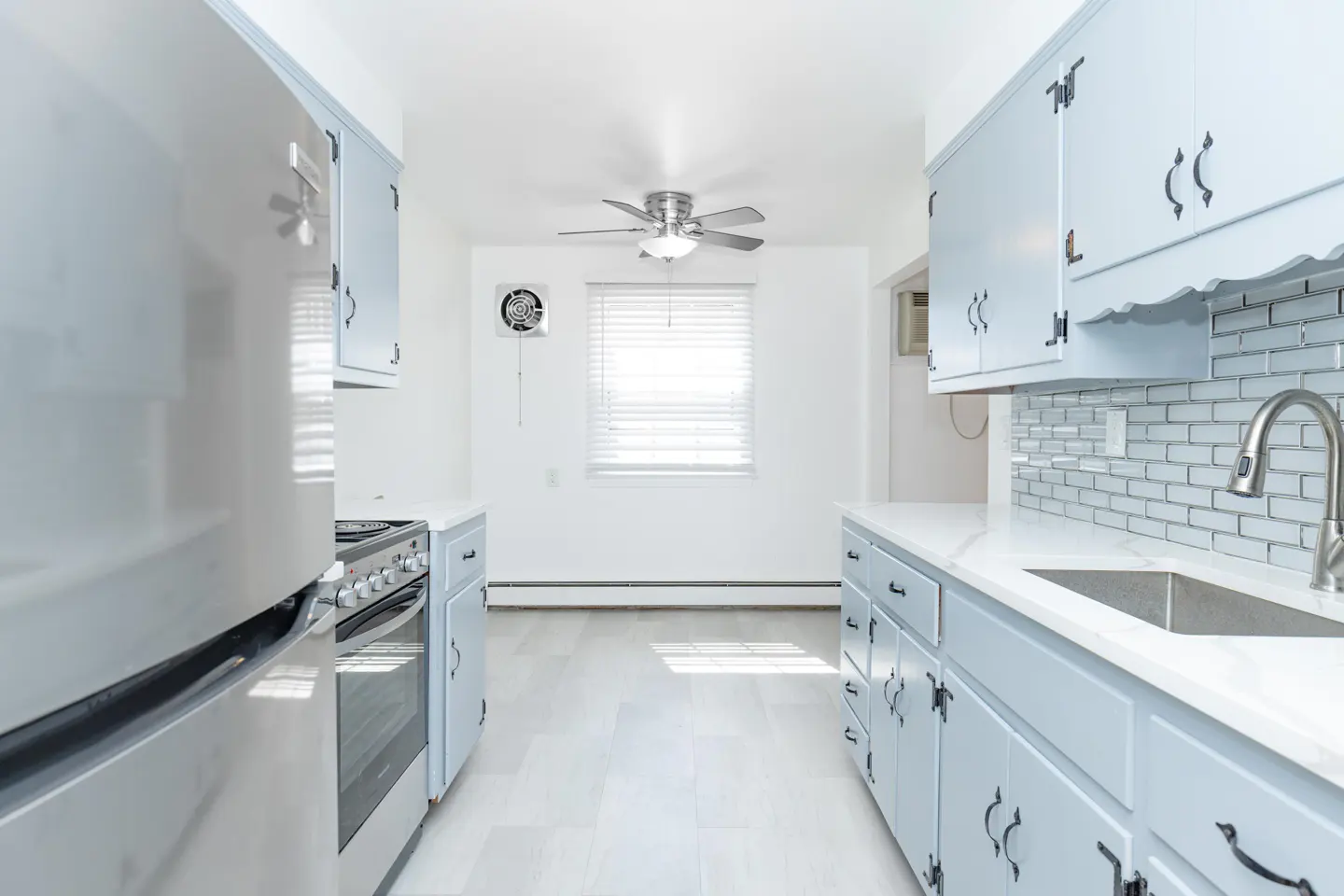 Bright kitchen with light blue cabinets, white countertops, and stainless steel appliances. A window with blinds is centered at the back.