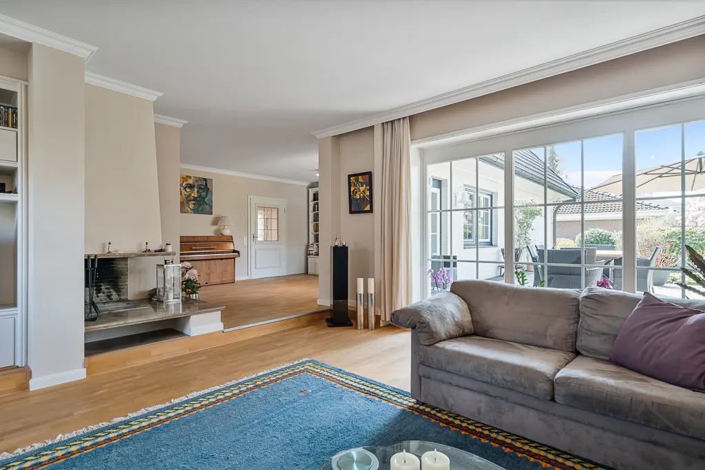 Living room with a gray sofa, blue rug, and a fireplace. A piano sits in the background, with large windows overlooking the patio.