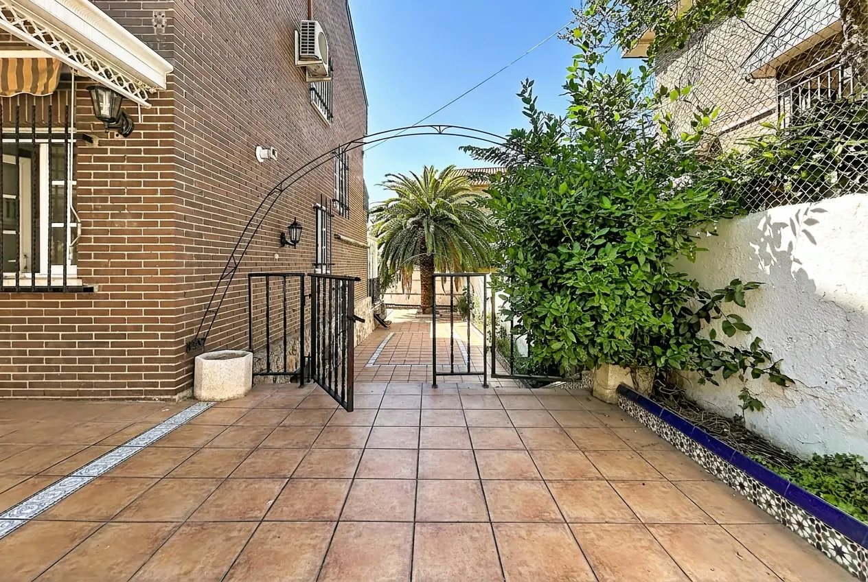 Exterior view of a brick house with a tiled patio and a wrought iron gate leading to a palm tree.