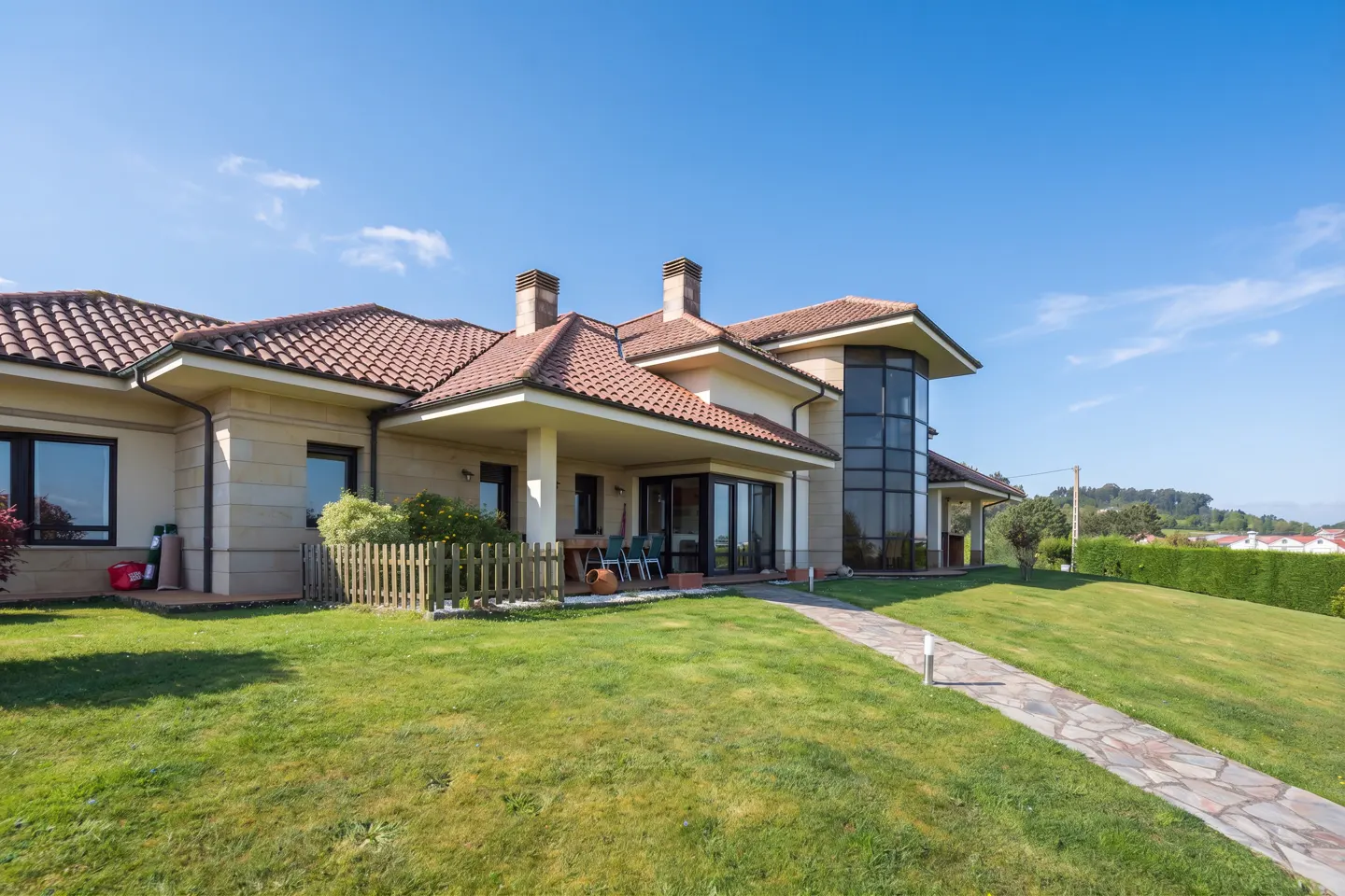 Exterior view of a beige house with a red tile roof, a glass tower, and a stone walkway on a sunny day.