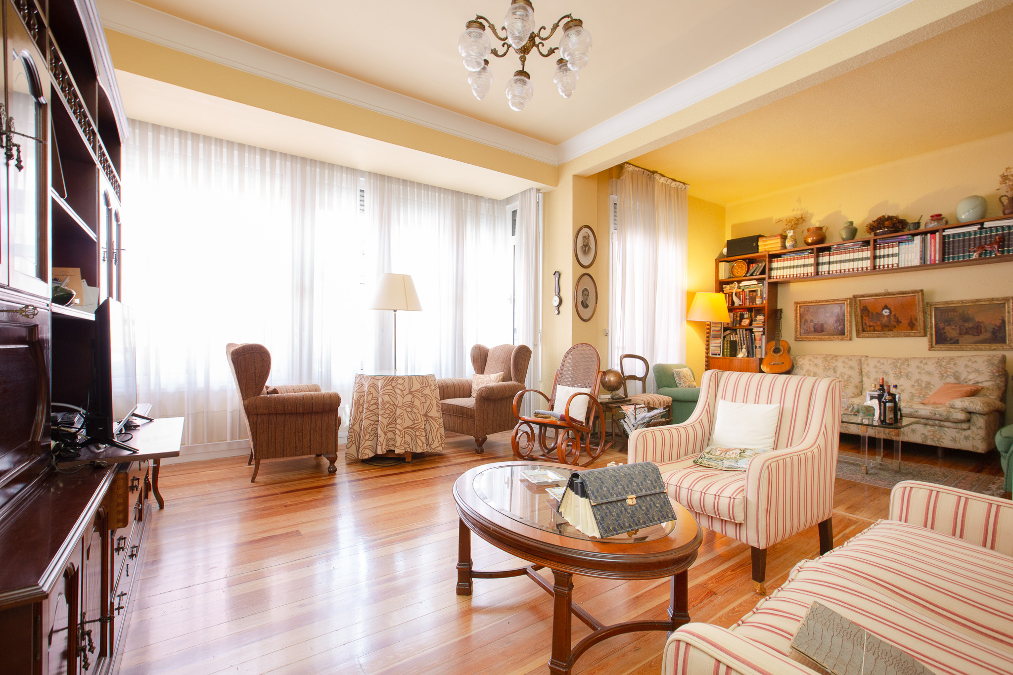 Bright living room with hardwood floors, striped armchairs, a wooden coffee table, and large windows with sheer curtains.