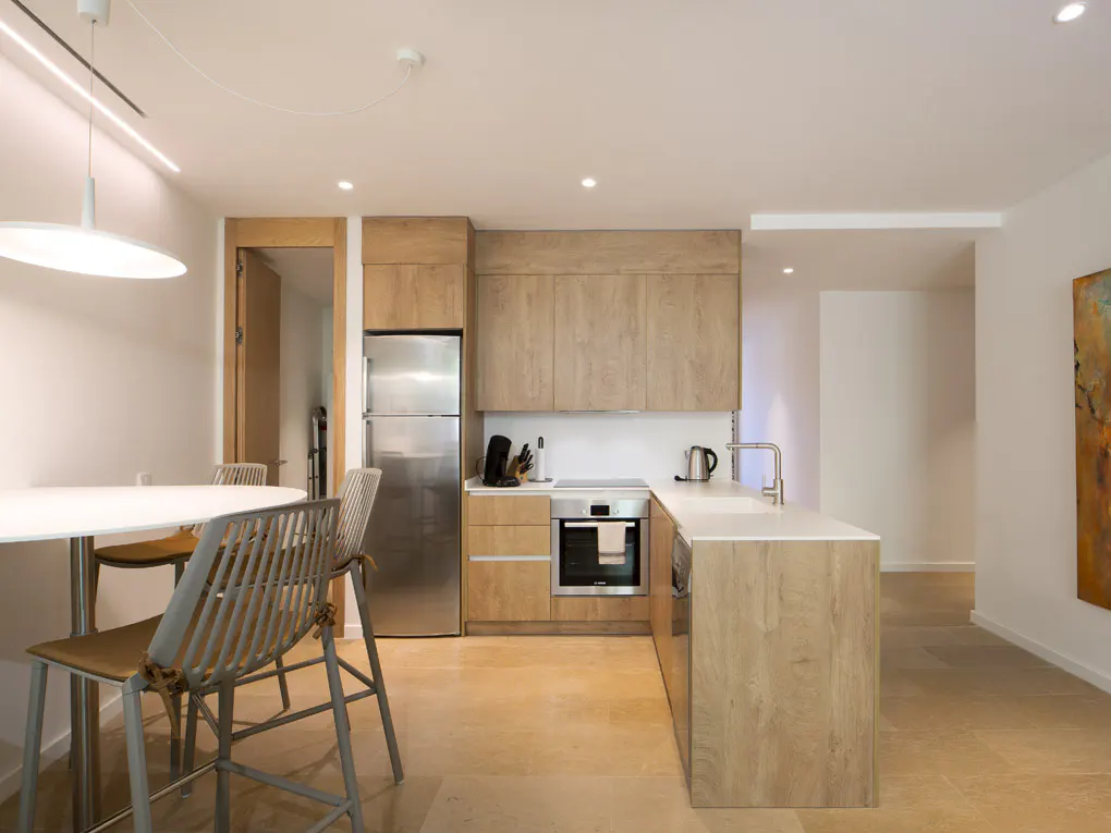 A bright, modern kitchen with light wood cabinets, stainless steel appliances, and a white countertop island. A round white table with chairs is nearby.