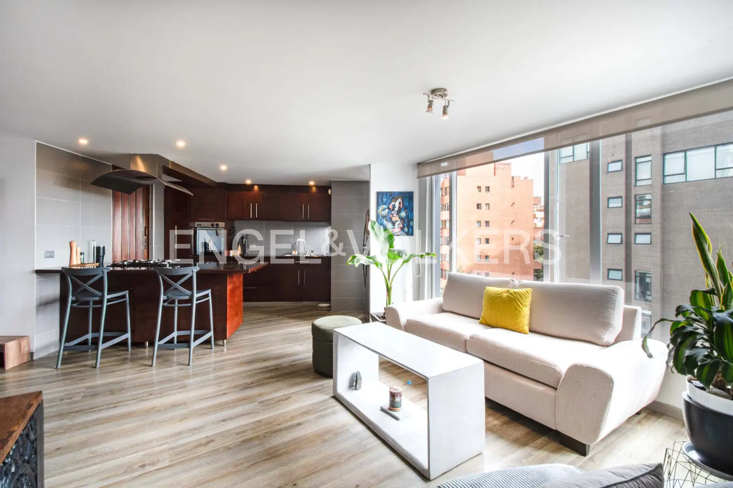 Open-concept apartment with wood floors, a kitchen island with bar stools, and a white sofa with a yellow pillow. Large windows show city views.