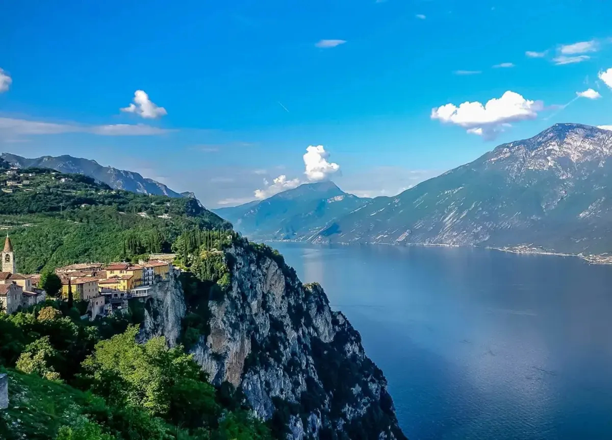 A scenic view of a village perched on a cliff overlooking a deep blue lake surrounded by mountains under a sunny sky. [AI-generated text]