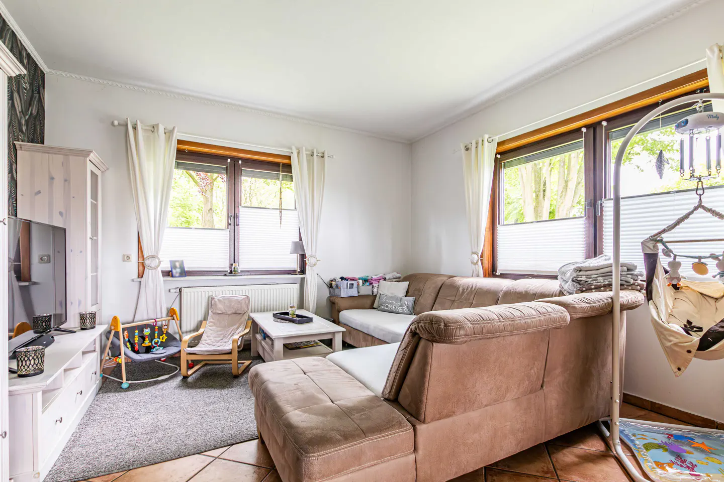 Living room with brown sectional sofa, white walls, and two windows with white curtains. A baby swing and play mat are visible.